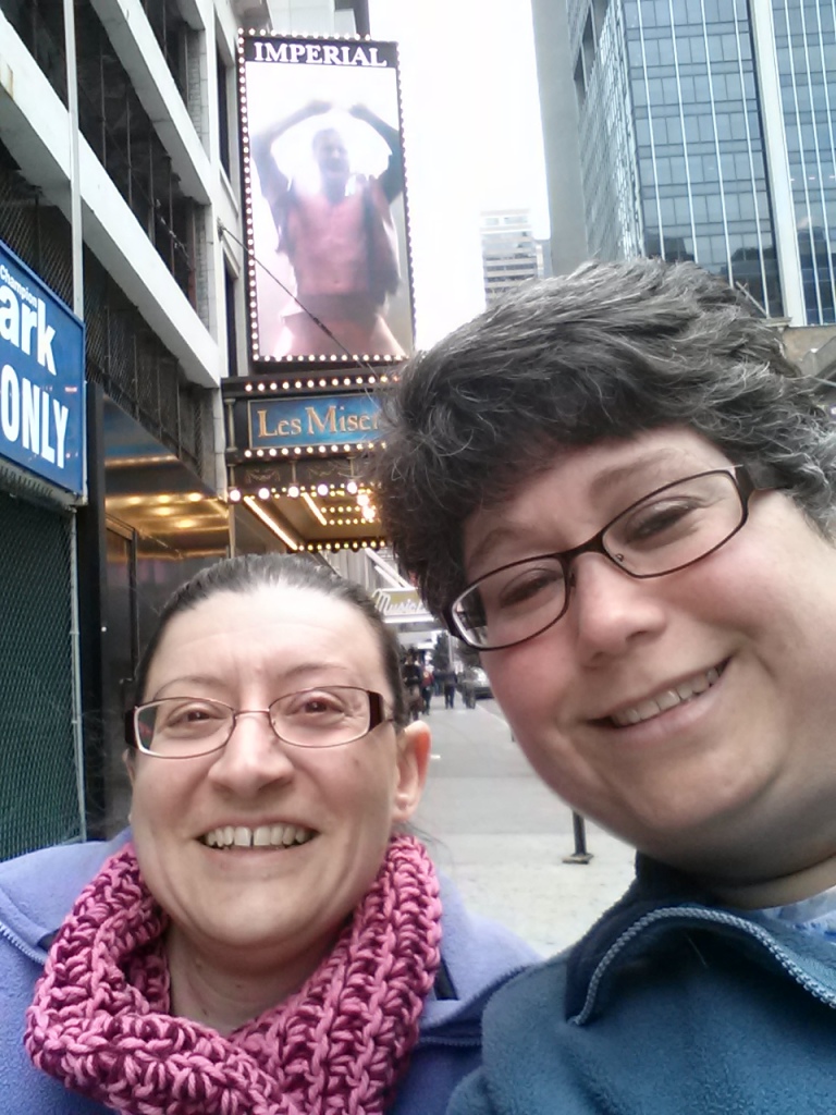 Photo of two women in front of a Broadway theater. Both have brown hair and are wearing glasses and smiling. They are posed in front of signs for Les Miserables.