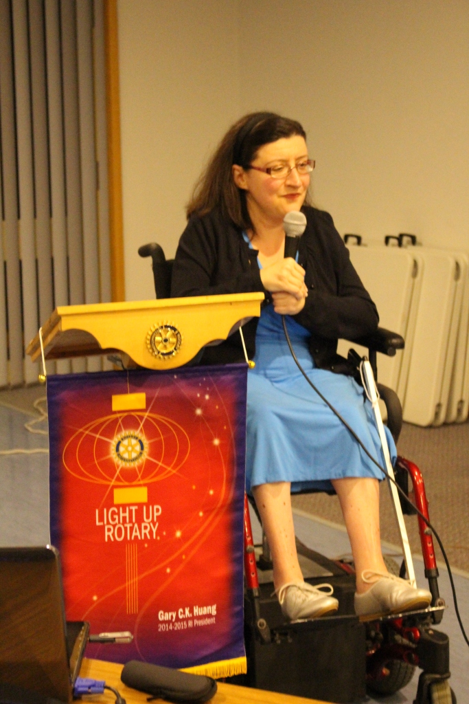 Photo of a woman in a wheelchair speaking into a microphone. She is seated behind a podium with a red banner which reads "Light Up Rotary."