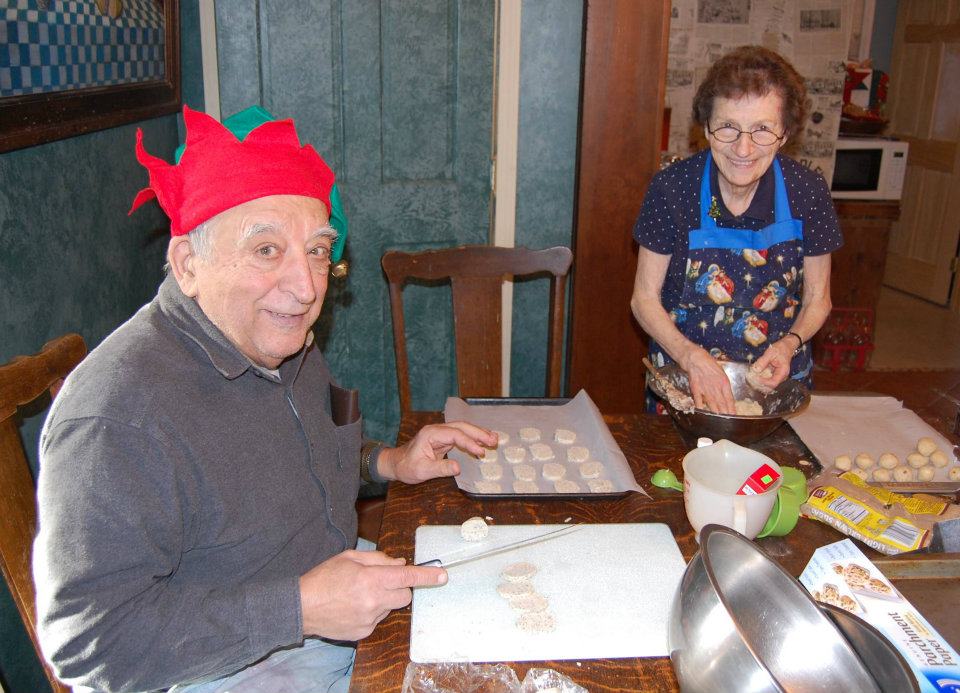 An elderly man wearing a red and green elf hat and an elderly woman wearing a blue apron smile at the camera. He is seated at a table and she is standing. They are making cookies.